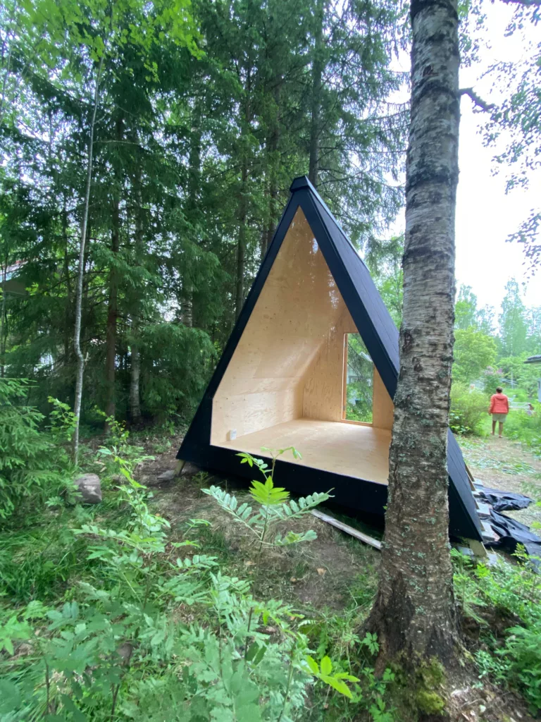 Kehto-mökki A-runkoinen puurakenne rakenteilla metsäiselle alueelle. Henkilö oranssissa paidassa seisoo taustalla lähellä puita.