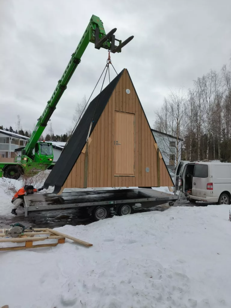 Kehto-mökki Pientä A-runkoista hyttiä nostetaan vihreällä nosturilla lumiselle maalle lähellä olevien työntekijöiden ja ajoneuvojen kanssa.